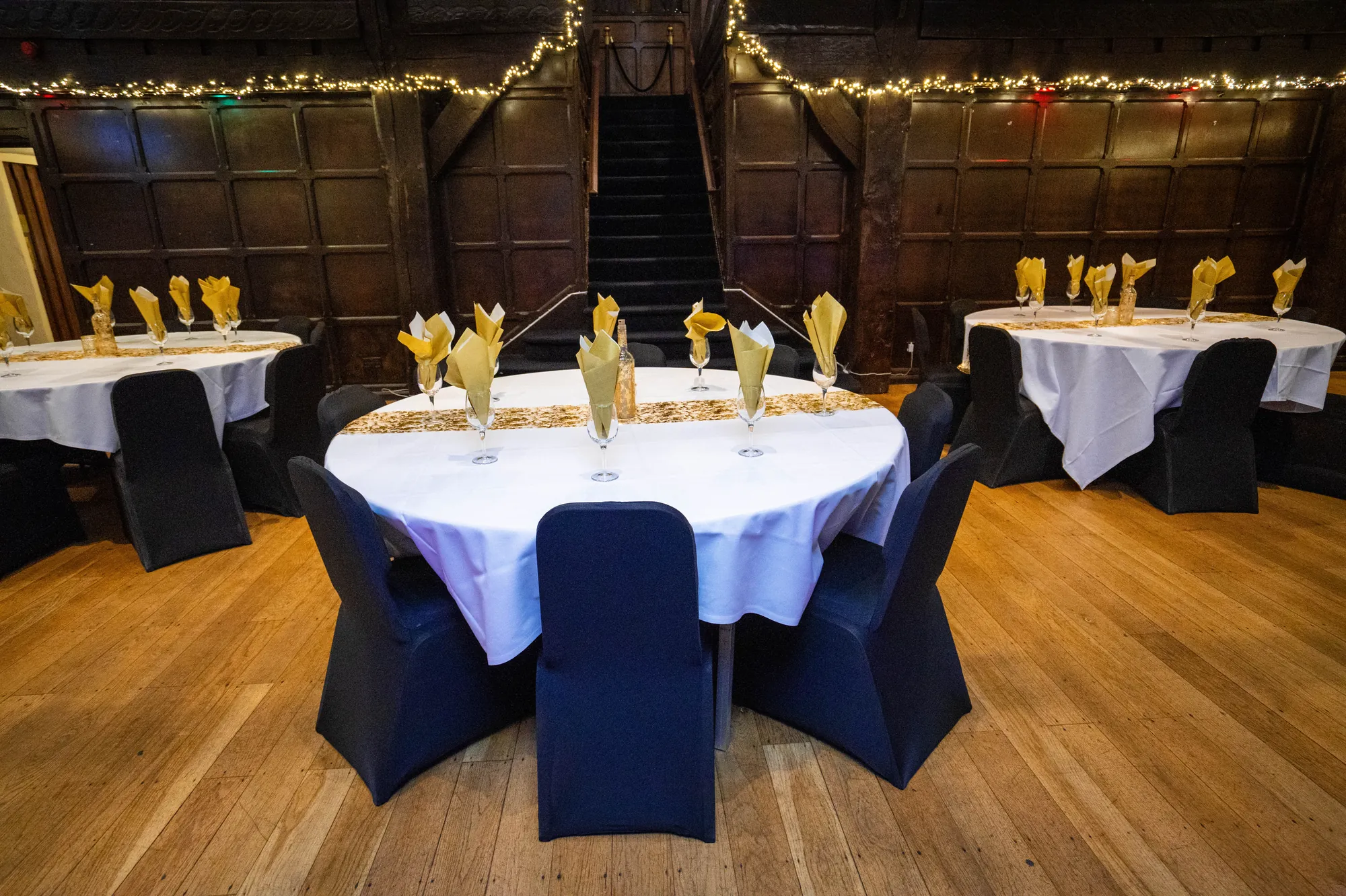 A round table in the Baronial Hall laid with white linen and gold, the panelled gallery and staircase behind