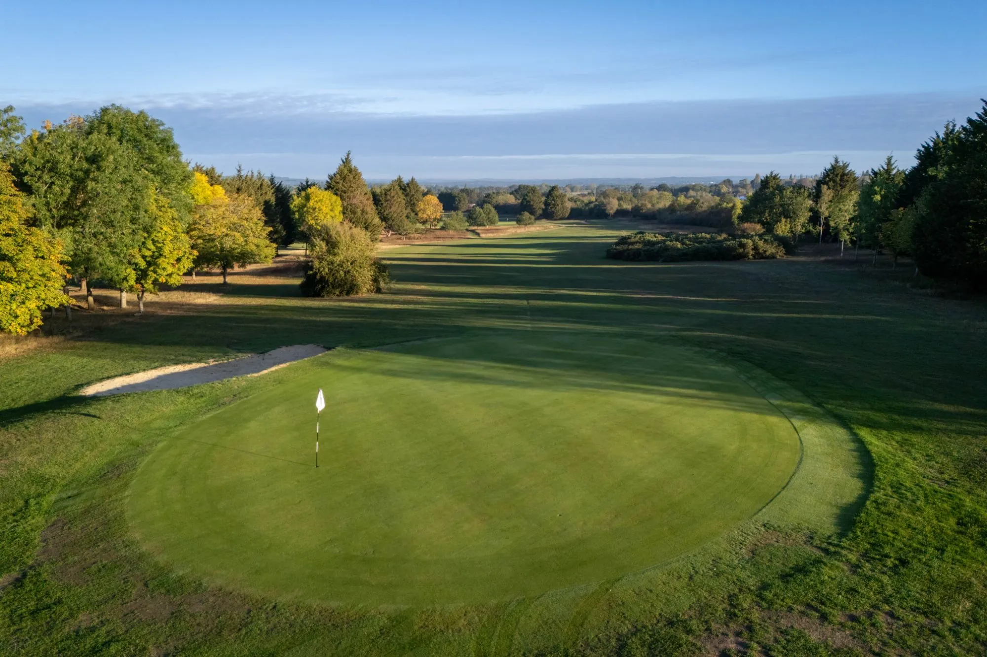 A green from the air with its flag and bunker, the fairway running away between autumn trees